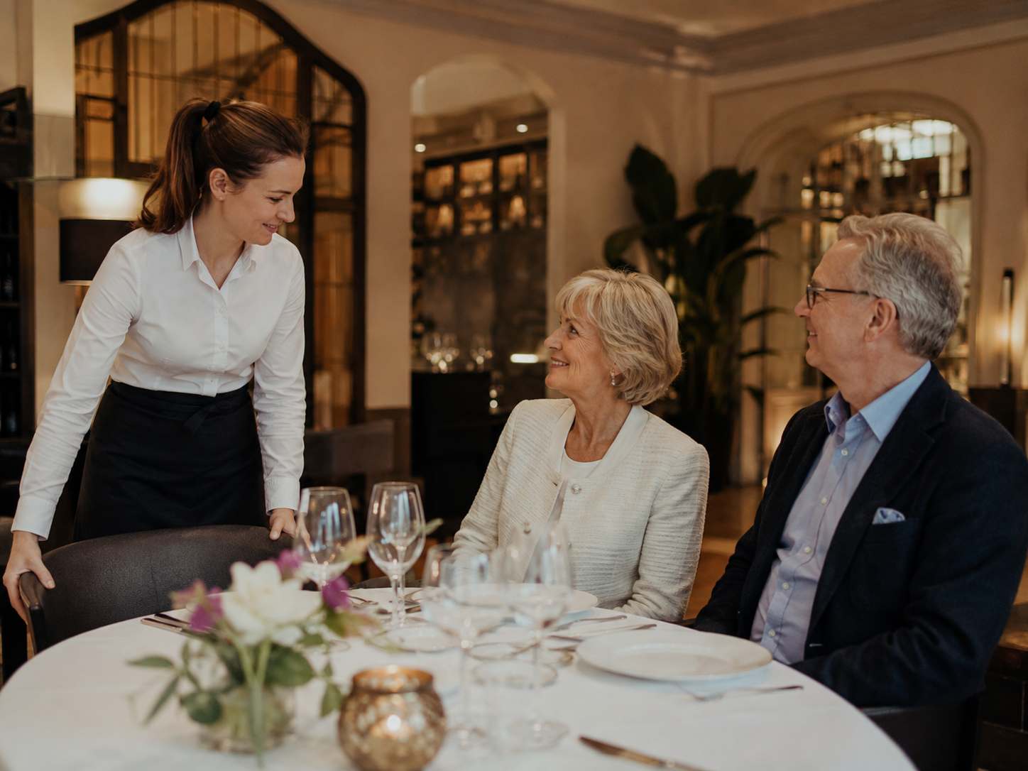 Servicekraft unterstützt ältere Gäste beim Platznehmen an einem gut erreichbaren Restauranttisch.