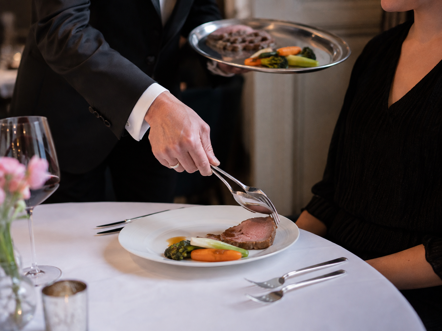 Servicekraft legt einer Gästin Fleisch und Gemüse mit Vorlegebesteck von einer Platte vor.